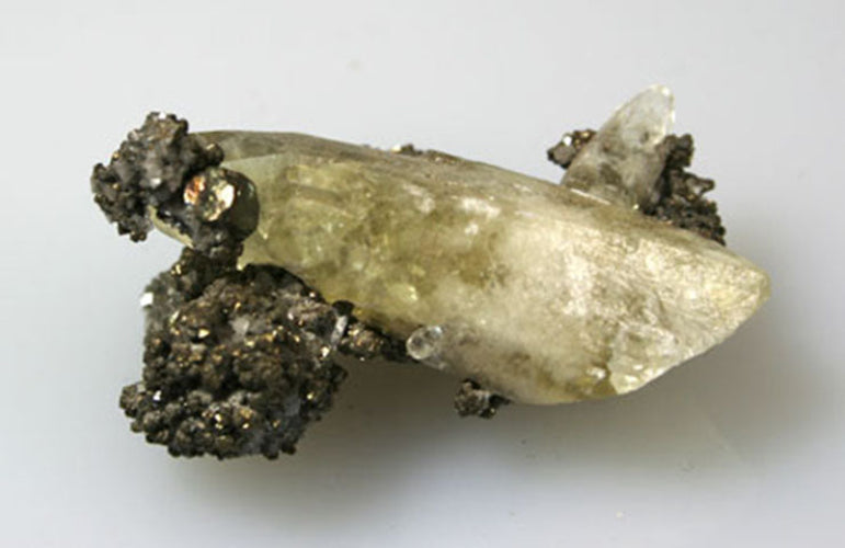 A natural mineral specimen featuring a translucent pale yellowish-green crystal cluster with dark metallic and silty matrix, photographed on a plain light background.