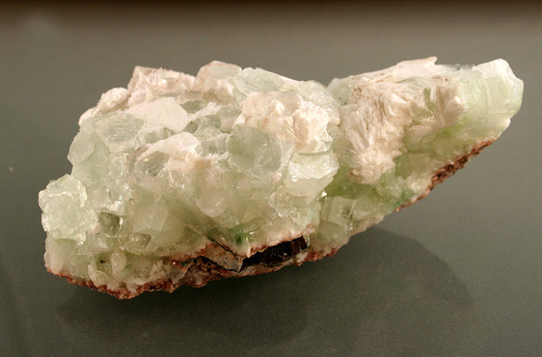 A natural mineral specimen with pale green and off-white crystalline formations on a dark gray reflective surface.