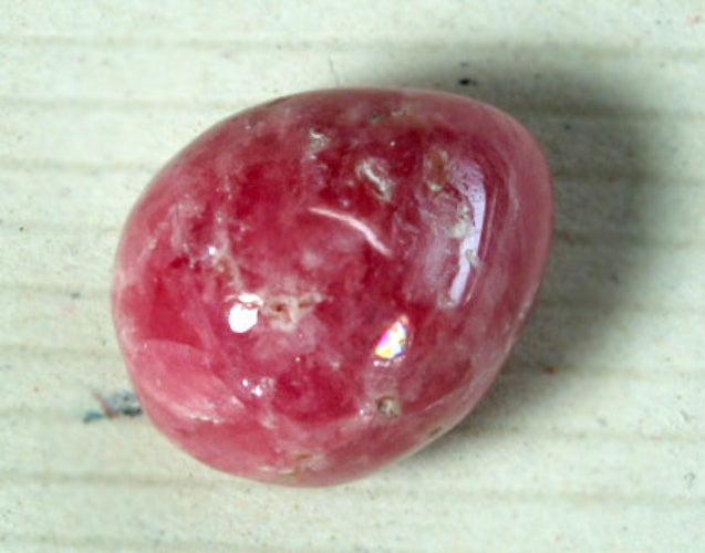 A polished, oval-shaped pink gemstone with natural inclusions and a glossy finish, resting on a light-colored textured surface.
