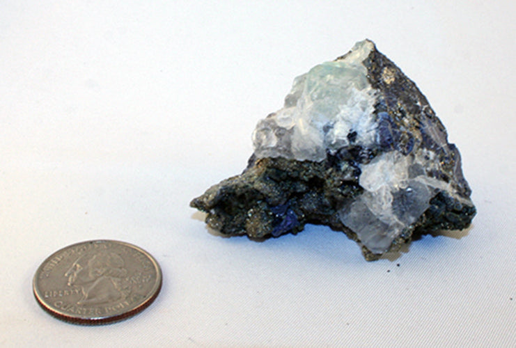A rough mineral specimen with pale blue and clear crystalline areas on a dark matrix, shown next to a US quarter coin for scale on a white background.