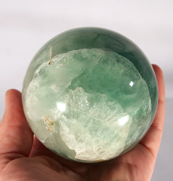 A hand holding a polished green crystal sphere with a natural cloudy interior, photographed against a plain light background.