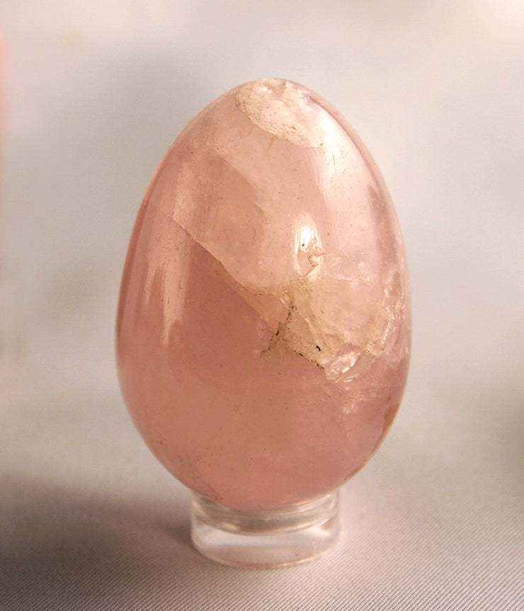 A polished pink rose quartz crystal egg on a small clear glass display stand, photographed against a neutral light gray background.